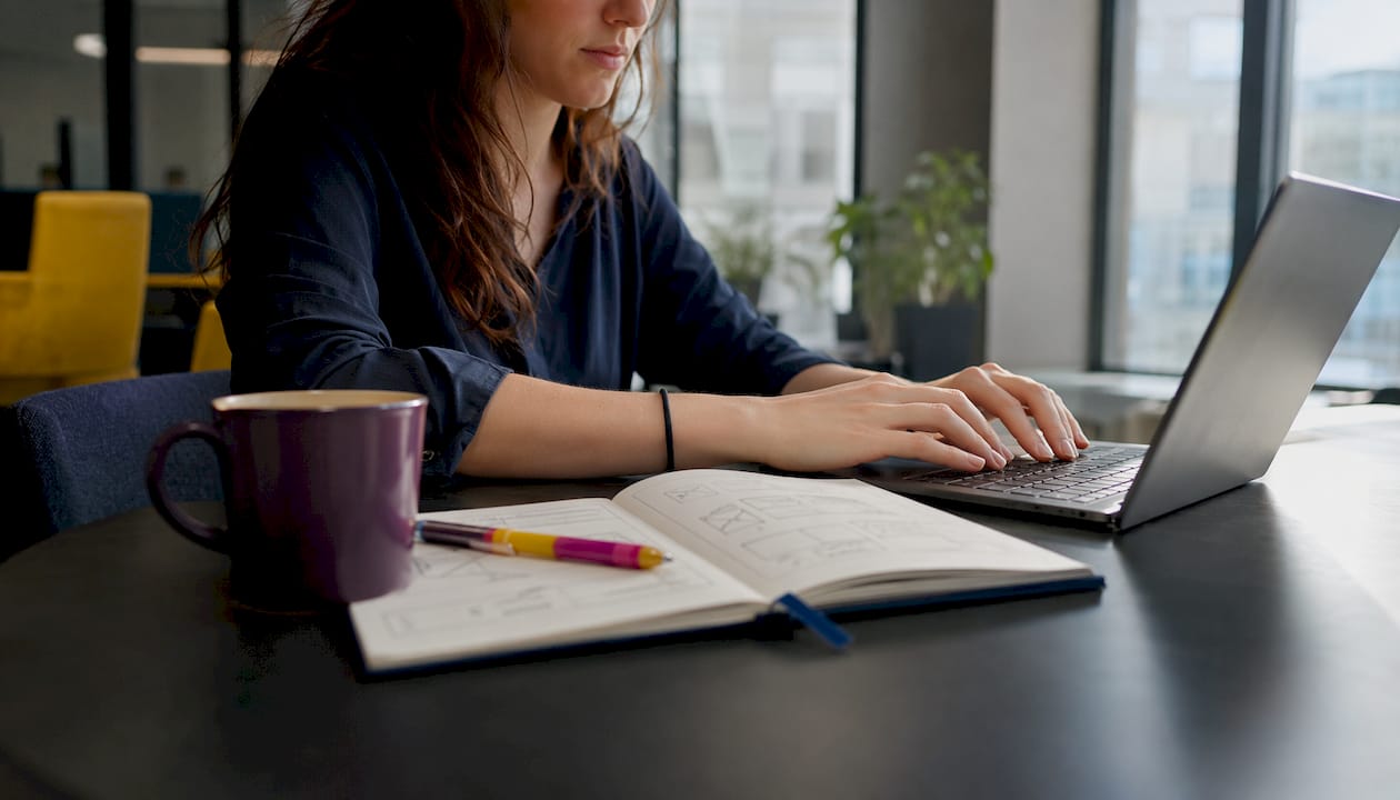 Mit einer Hand am Laptop, die andere greift zum Kaffeebecher – daneben liegt das geöffnete Notizbuch.